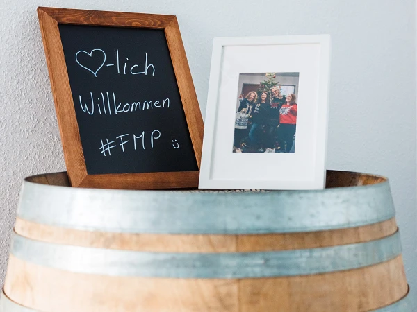 Ein Holzfass dient als Ablage für ein kleines, gerahmtes Foto und eine Tafel mit der handgeschriebenen Begrüßung „Herzlich Willkommen #FMP“.