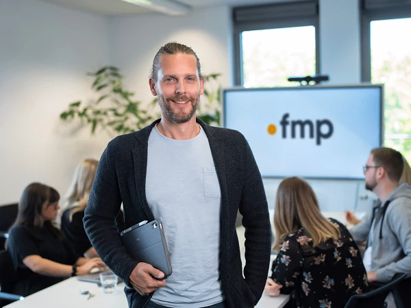Ein Mann mit langen Haaren und einem breiten Lächeln steht in einem modernen Büro und hält ein Tablet in der Hand, während im Hintergrund eine Gruppe von Menschen an einem Tisch arbeitet und ein Bildschirm mit dem Logo „fmp“ zu sehen ist.