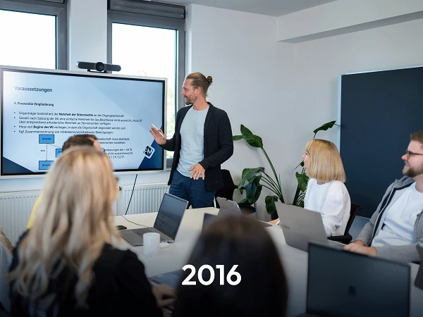 Ein männlicher Präsentator steht vor einem Bildschirm in einem modernen Besprechungsraum und erklärt die Inhalte einer Präsentation. Die Gruppe von fünf Personen, die auf Stühlen sitzt, schaut aufmerksam zu. Auf dem Bildschirm sind die Worte "Voraussetzungen" und einige grafische Elemente zu sehen, die offensichtlich einen Überblick über die Themen der Präsentation geben. Im Vordergrund sind die Köpfe der Zuhörer teilweise sichtbar.