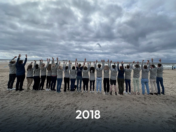 Eine Gruppe von Menschen steht am Strand, mit dem Gesicht zum Meer, und hebt beide Arme in die Luft. Die Personen tragen helle Sweatshirts und eine Wolkendecke ist am Himmel sichtbar. Unter dem Motiv steht die Jahreszahl "2018".