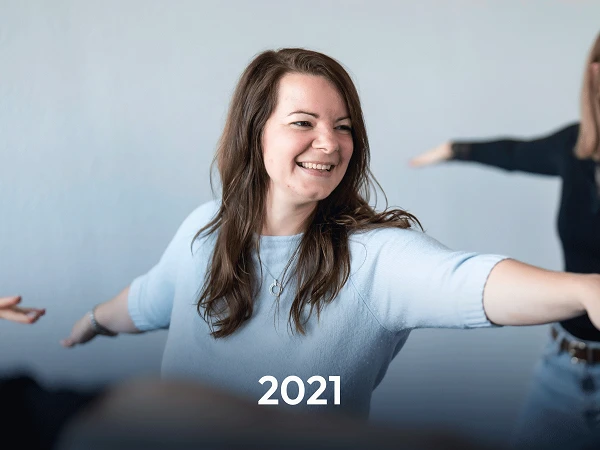 Eine lächelnde Frau in einem hellen Pullover mit langen Haaren stellt eine einladende Geste dar. Im Hintergrund sind verschwommen weitere Personen zu sehen, die ebenfalls eine ähnliche Pose einnehmen. Unter dem Bild steht die Jahreszahl „2021“.