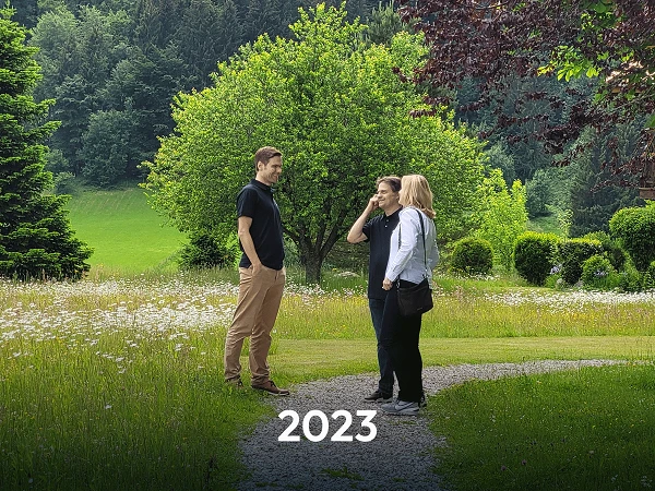 Drei Personen stehen auf einem Weg in einem grünen Garten, umgeben von Bäumen und Blumen, während sie miteinander sprechen. Im Vordergrund steht die Jahreszahl "2023".