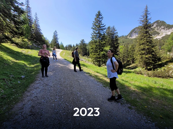 Fünf Personen stehen auf einem Schotterweg in einer bewaldeten Gebirgslandschaft, umgeben von grünen Bäumen und Bergen. Am unteren Bildrand ist die Jahreszahl „2023“ in großer Schrift zu sehen.