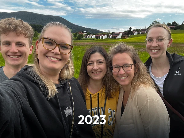 Eine Gruppe von fünf Personen macht ein Selfie vor einer grünen Wiesenlandschaft, umgeben von Bergen und einem Wohngebiet, mit dem Jahr „2023“ in der Mitte des Bildes.