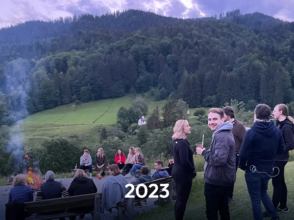 Eine Gruppe von Menschen versammelt sich um ein Feuer in der Abenddämmerung, umgeben von einer malerischen Berglandschaft. Im Vordergrund steht ein Mann, der in die Kamera lächelt, während andere Personen mit Getränken plaudern. Im unteren Bereich des Bildes ist das Jahr „2023“ in großer Schrift zu sehen.
