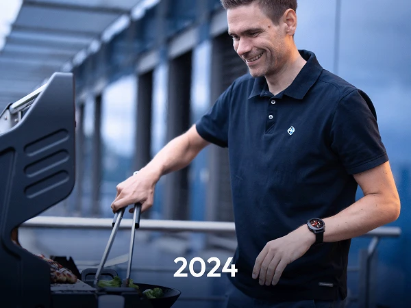 Ein Mann in einem schwarzen Polohemd steht an einem Grill und wendet mit Zangen Fleisch und Gemüse, während er lächelt. Im Vordergrund ist die Zahl „2024“ zu sehen.
