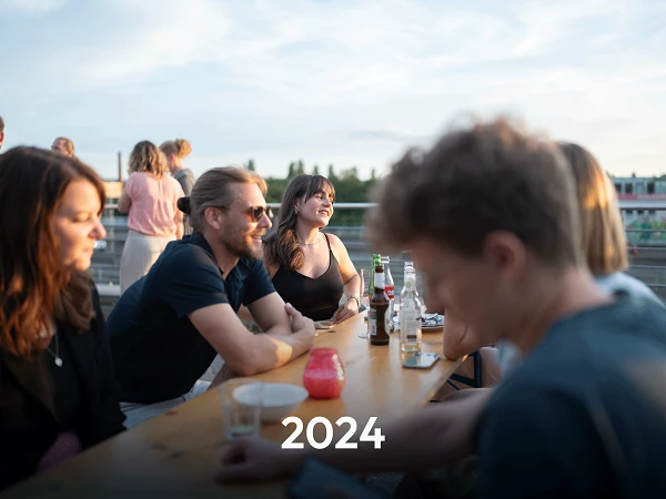 Eine Gruppe von Menschen sitzt an einem Holztisch auf einer Terrasse, während die Sonne untergeht. Einige Getränke stehen auf dem Tisch, und im Vordergrund ist das Jahr „2024“ in großen Zahlen abgebildet.