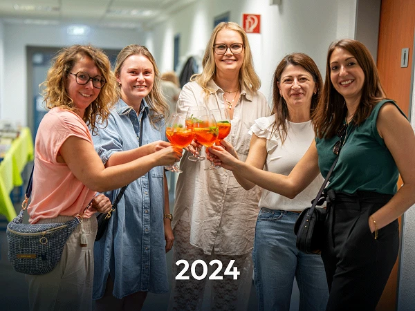 Eine Gruppe von fünf Frauen steht in einem Flur und hält Gläser mit bunten Cocktails in der Hand, während sie anstoßen. Im Hintergrund sind Teilbereiche eines Veranstaltungssaals sichtbar. Am unteren Bildrand steht die Zahl „2024“.