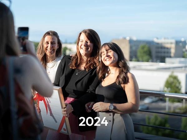 Drei Frauen posieren lächelnd für ein Foto auf einer Terrasse, während eine vierte Person sie mit einer Kamera aufnimmt. Im Vordergrund steht das Jahr „2024“ in großer Schrift.