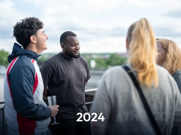 Eine Gruppe von vier Personen in casual Kleidung unterhält sich auf einem Balkon mit Blick auf eine grüne Landschaft und einen bewölkten Himmel. In der Mitte steht ein Mann mit einem freundlichen Lächeln. Der Text „2024“ steht prominent in der Mitte des Bildes.