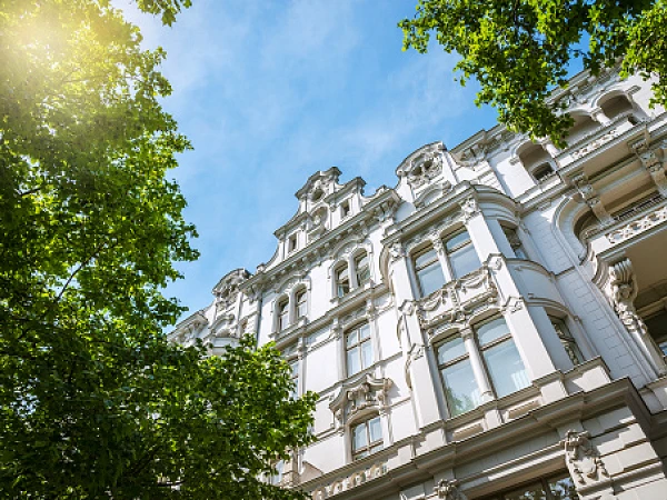 Die Fassade eines historischen Gebäudes mit dekorativen Elementen ist von grünen Baumblättern umgeben, während der Himmel blau und lichtdurchflutet ist.