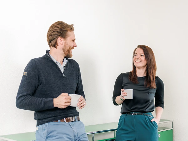 Ein Mann und eine Frau stehen lächelnd an einem grünen Sideboard und unterhalten sich, während sie Kaffeetassen in den Händen halten. Der Mann trägt einen dunkelblauen Pullover und eine hellblaue Hose, die Frau hat ein schwarzes Oberteil und eine grüne Hose an.
