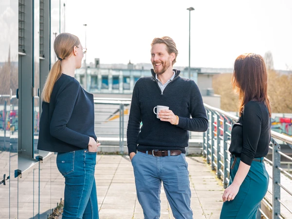 Drei Personen stehen auf einer sonnigen Terrasse und unterhalten sich, während eine Person eine Tasse hält. Die Frau links trägt einen schwarzen Pullover und jeans, die Frau rechts eine schwarze Bluse und grüne Hose, und der Mann in der Mitte hat einen grauen Pullover und trägt eine lockere Hose.
