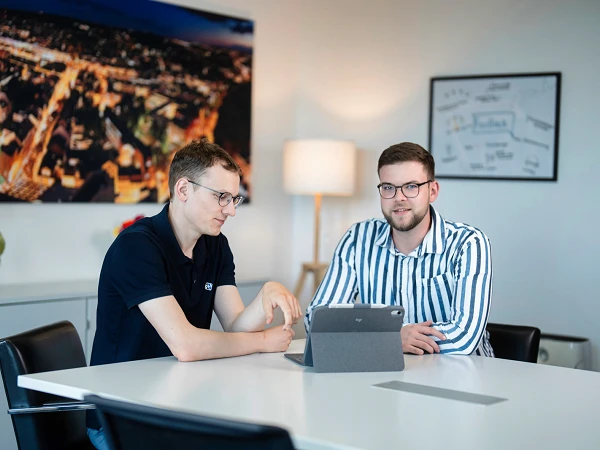 Zwei Männer sitzen an einem Tisch in einem modernen Büroraum und schauen auf ein Tablet. Im Hintergrund ist eine große Stadtansicht an der Wand zu sehen.