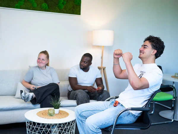Eine Gruppe von drei Personen sitzt in einem modernen Raum. Eine Person sitzt auf einem Stuhl und zeigt mit beiden Händen eine triumphierende Geste, während die anderen beiden entspannt auf einer Couch sitzen. Auf dem Tisch in der Mitte steht eine kleine Pflanze und eine Kerze.