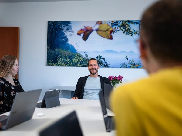 Eine Gruppe von fünf Personen sitzt an einem Tisch in einem Besprechungsraum; ein Mann in der Mitte lächelt, während er mit den anderen spricht. Im Hintergrund hängt ein Landschaftsbild mit Bäumen und Bergen.