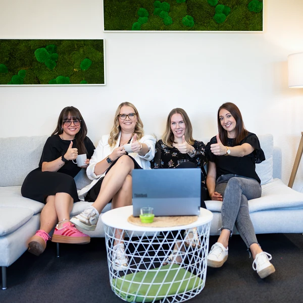 Vier Frauen sitzen auf einem hellen Sofa in einem modernen Büro. Sie zeigen Daumen nach oben und lächeln, während ein Laptop auf dem Tisch vor ihnen steht. Im Hintergrund sind grüne Wanddekorationen und eine Stehlampe zu sehen.