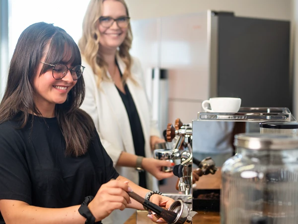 Zwei Frauen bereiten Kaffee an einer modernen Kaffeebar in einem hellen Raum zu, während sie lächeln und miteinander sprechen.