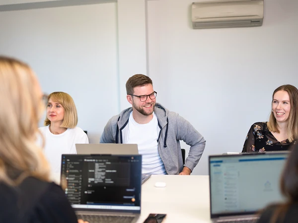 Eine Gruppe von vier Personen sitzt an einem Tisch in einem modernen Besprechungsraum, während sie miteinander sprechen. Alle haben Laptops vor sich, und einige Menschen lächeln. Im Vordergrund sind ihre Laptops und ein Smartphone sichtbar.