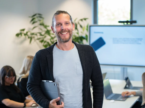 Ein Mann steht lächelnd in einem modernen Besprechungsraum, während im Hintergrund mehrere Personen an einem Tisch arbeiten. Der Mann hält eine Dokumentenmappe in der Hand, und auf einem Bildschirm wird ein Präsentationsinhalt angezeigt.