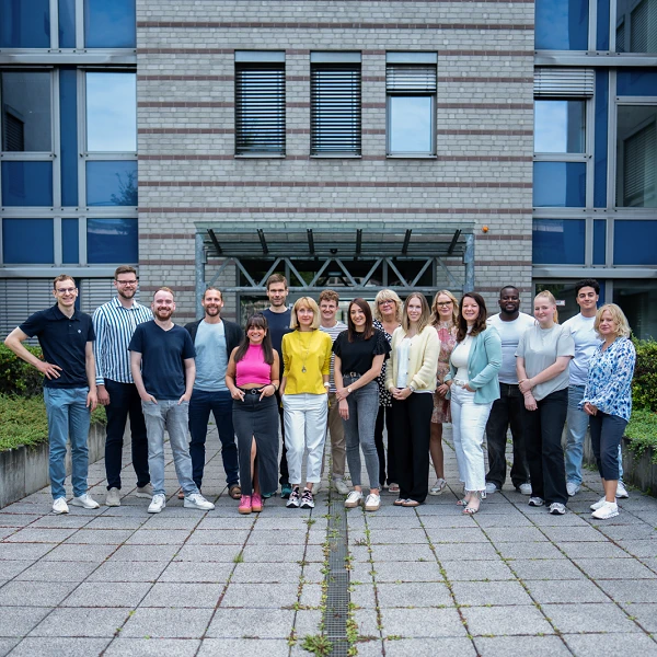 Eine Gruppe von 17 Personen steht lächelnd in einem Innenhof zwischen modernen Gebäuden, umgeben von grüner Bepflanzung und Steinen. Die Personen tragen verschiedene Freizeitoutfits und posieren freundlich für das Foto.
