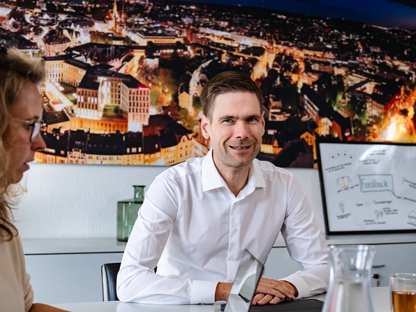 Ein Mann in einem weißen Hemd sitzt an einem Tisch in einem Besprechungsraum und lächelt, während um ihn herum mehrere Personen mit Laptops und einem Tablet beschäftigt sind. Im Hintergrund ist ein beleuchtetes Stadtbild an der Wand zu sehen.
