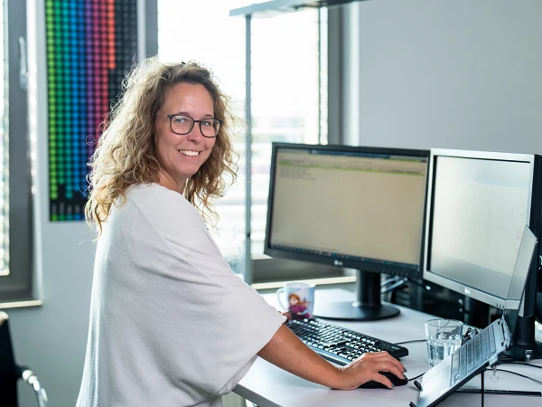 Eine lächelnde Frau mit lockigem Haar sitzt an einem Schreibtisch und arbeitet an zwei Monitoren, während sie auf die Kamera blickt. Die Umgebung wirkt modern und hell, mit einer Glaswasserflasche und einem Ständer für den Laptop auf dem Tisch.