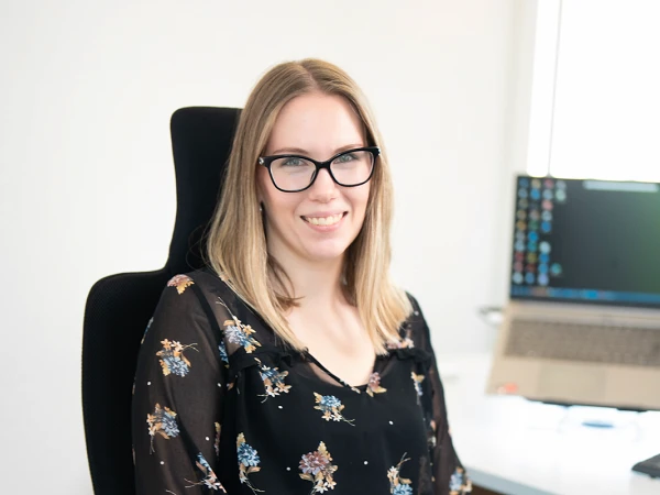 Eine Frau mit Brille sitzt lächelnd in einem Bürostuhl in einem gut beleuchteten Büro, umgeben von einem Computer und einem Schreibtisch. Sie trägt ein schwarzes Oberteil mit floralem Muster und hat lange, glatte Haare.