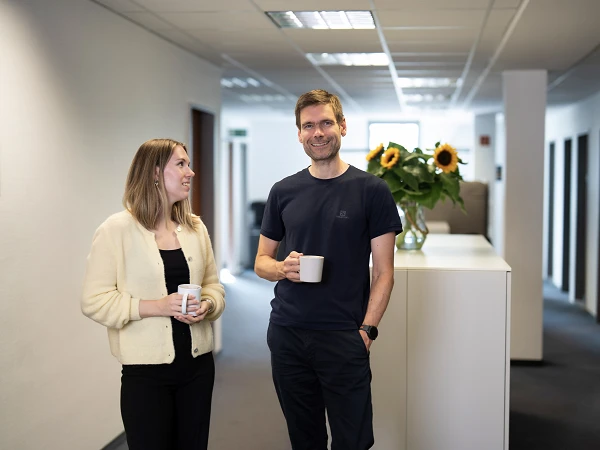 Ein Mann und eine Frau stehen in einem modernen Büroflur, beide halten eine Tasse in der Hand. Die Frau schaut den Mann an und lächelt, während im Hintergrund ein großer Blumenstrauß mit Sonnenblumen zu sehen ist.