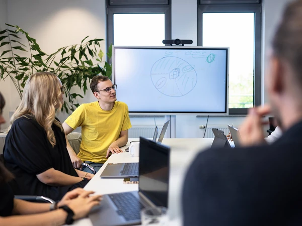 Eine Gruppe von Menschen sitzt an einem Konferenztisch, während ein junger Mann in gelbem T-Shirt auf einen Bildschirm zeigt, auf dem eine Zeichnung zu sehen ist. Im Hintergrund sind Pflanzen und Fenster erkennbar.