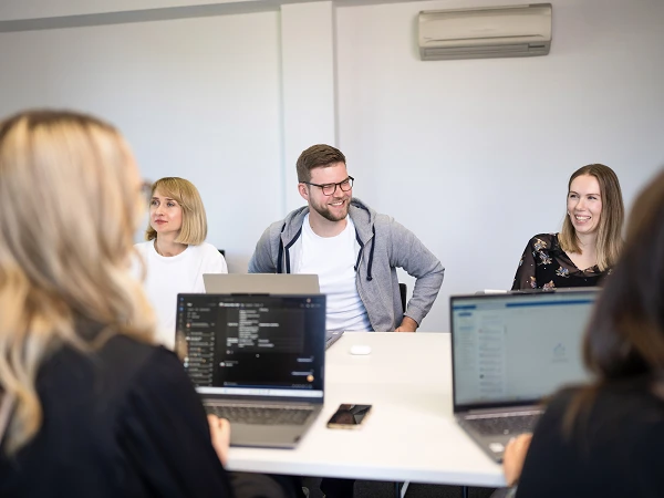 Eine Gruppe von vier Personen sitzt an einem Tisch in einem modernen Büro, während sie sich unterhalten. Zwei Laptops sind auf dem Tisch sichtbar, und der Mann in der Mitte trägt eine Brille sowie ein graues Sweatshirt.