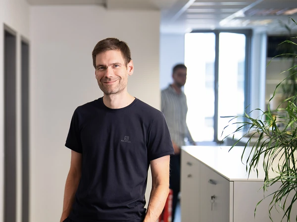 Zwei Personen stehen in einem modernen Büroraum, während sie sich unterhalten; im Hintergrund sind weitere Büros und ein Pflanzenarrangement sichtbar.