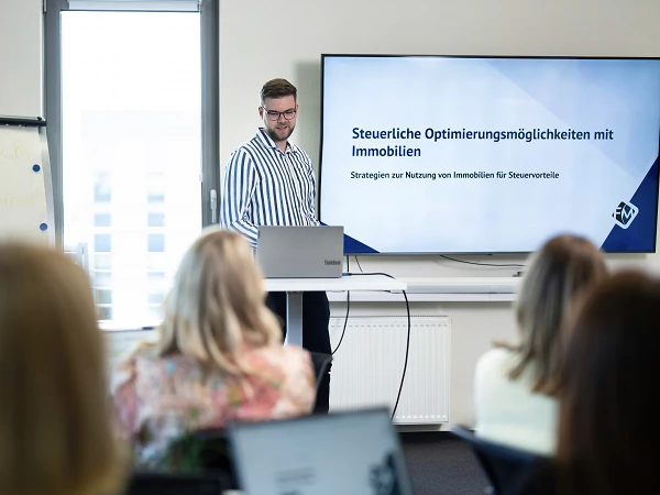 Ein Mann präsentiert vor einer Gruppe von Zuhörern in einem Seminarraum, während im Hintergrund eine Bildschirmpräsentation mit dem Titel „Steuerliche Optimierungsmöglichkeiten mit Immobilien“ zu sehen ist. Der Mann spricht über Strategien zur Nutzung von Immobilien für Steuerzwecke. Die Zuhörer sind teilweise mit Laptops beschäftigt.