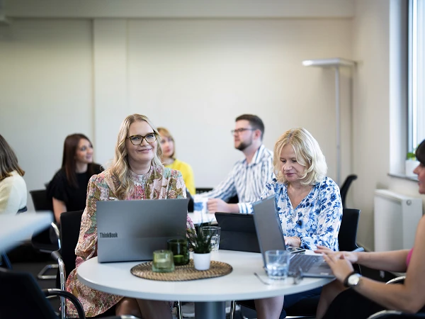 Eine Gruppe von fünf Personen in einem modernen Büroraum sitzt an einem runden Tisch mit Laptops, während sie miteinander kommunizieren und arbeiten. Es gibt Pflanzen und Gläser mit Wasser auf dem Tisch.