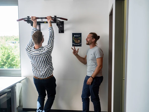Ein Mann hängt an einer Trainingsstange an der Wand, während ein anderer Mann daneben steht und ihn anfeuert oder mit ihm spricht. Im Hintergrund ist ein Fenster mit Blick ins Grüne sichtbar.