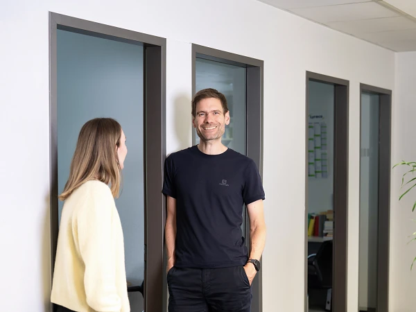 Ein Mann steht lächelnd in einem modernen Büroflur, während eine Frau ihm gegenübersteht und mit ihm spricht. Im Hintergrund sind Bürotüren und Einrichtungen sichtbar.