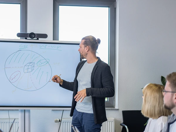 Ein Mann präsentiert an einem Whiteboard in einem modernen Besprechungsraum, während fünf Personen sitzend, mit Laptops vor sich, aufmerksam zuhören. Einige Pflanzen sind im Raum sichtbar, und es gibt große Fenster, die Tageslicht hereinlassen.