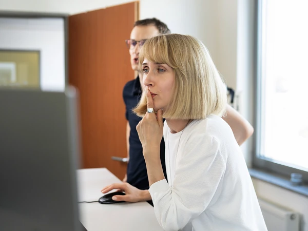 Eine Frau mit blonden, schulterlangen Haaren und einem weißen Oberteil betrachtet nachdenklich einen Computerbildschirm, während ein Mann im Hintergrund ebenfalls auf den Bildschirm schaut. Der Raum ist hell und modern eingerichtet.
