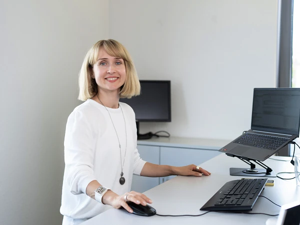 Eine junge Frau mit blonden Haaren steht an einem modernen Schreibtisch und lächelt, während sie mit der Maus an ihrem Computer arbeitet. Auf dem Schreibtisch befinden sich mehrere Monitore und ein Laptop. Die Büroumgebung ist hell und freundlich.