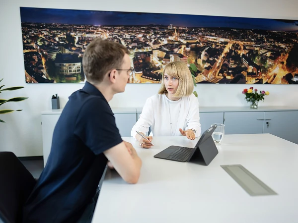 Eine Frau mit blonden Haaren und einem weißen Oberteil sitzt an einem Tisch und spricht mit einem Mann in einem schwarzen T-Shirt; im Hintergrund ist eine städtische Skyline bei Nacht zu sehen. Auf dem Tisch steht ein Glas Wasser und ein Tablet.
