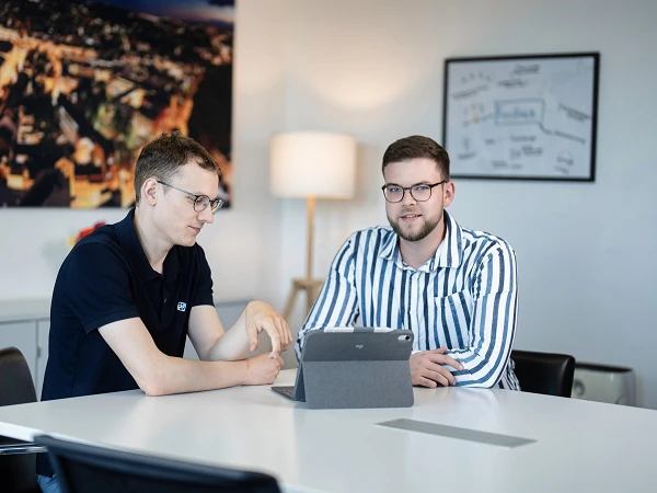 Zwei Männer sitzen an einem Tisch in einem modern eingerichteten Büro, während sie auf ein Tablet schauen. Im Hintergrund ist ein großes Stadtbild und ein Flipchart zu sehen.