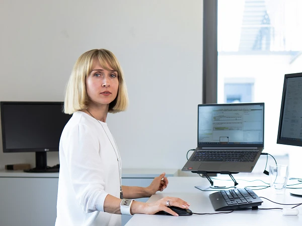 Eine Frau mit blonden, schulterlangen Haaren steht an einem höhenverstellbaren Schreibtisch in einem modernen Büro, während sie an einem Computer arbeitet. Der Raum ist hell und minimalistisch eingerichtet, mit mehreren Bildschirmen und Büroausstattung im Hintergrund.