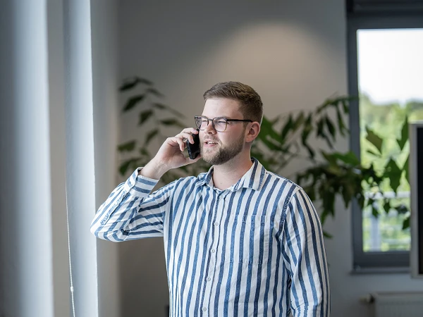 Ein Mann mit Brille trägt ein gestreiftes Hemd und telefoniert in einem modernen Büro. Im Hintergrund sind große Fenster und Pflanzen zu sehen.