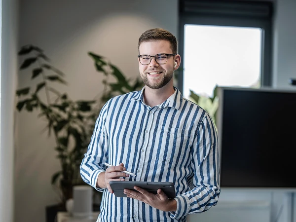 Ein junger Mann mit Brille steht in einem modernen Büro und hält ein Tablet in der Hand, während er in die Kamera lächelt. Im Hintergrund sind Pflanzen und ein Computerbildschirm zu sehen.