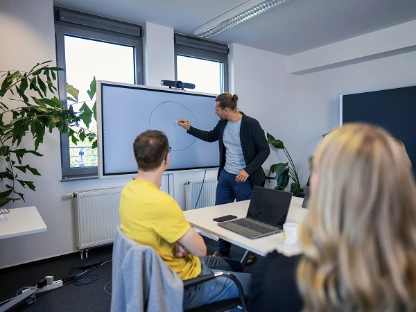 Ein Mann zeichnet einen Kreis auf einem digitalen Bildschirm während einer Präsentation in einem modernen Konferenzraum. Im Vordergrund sitzen mehrere Personen, die ihm aufmerksam zusehen. Pflanzen und Fenster mit Tageslicht sind im Hintergrund sichtbar.