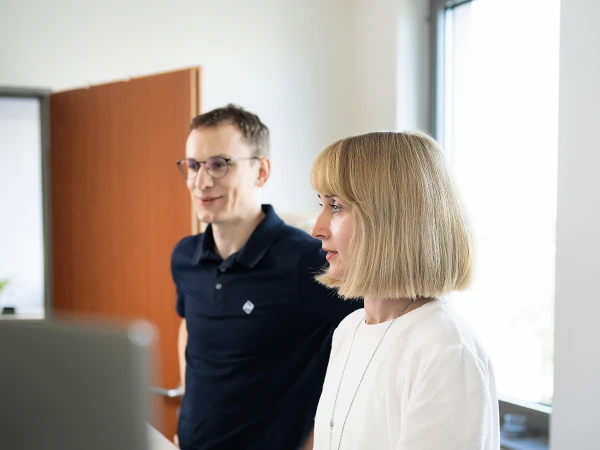 Ein Mann und eine Frau stehen nebeneinander und schauen auf einen Computerbildschirm. Der Mann trägt ein schwarzes Polohemd und hat eine Brille auf, während die Frau ein weißes Oberteil trägt und kurze, blonde Haare hat. Im Hintergrund sind Fenster und eine Holztür zu sehen.