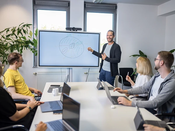 Ein Mann steht vor einem Bildschirm in einem Besprechungsraum und erklärt eine Grafik, während mehrere Personen an einem Tisch mit Laptops aufmerksam zuhören. Pflanzen sind im Hintergrund sichtbar.