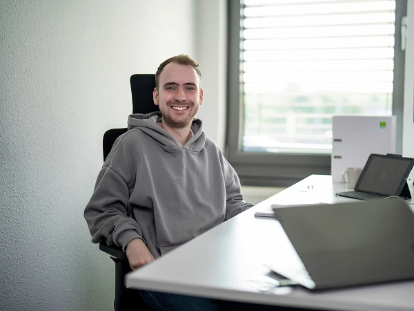 Ein junger Mann mit einem grauen Hoodie sitzt lächelnd an einem Schreibtisch, der mit einem Laptop, einem Notizbuch und einem Monitor ausgestattet ist. Hinter ihm ist ein Fenster mit Jalousien, das Tageslicht in den Raum lässt.
