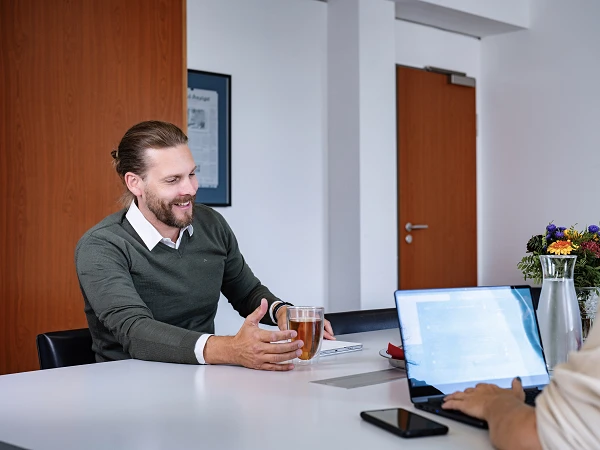 Ein Mann mit leichtem Bart sitzt an einem Tisch und hält ein Glas mit Getränk in der Hand, während er mit einer Frau spricht, die an einem Laptop arbeitet. Im Hintergrund sind Blumen und eine Wasserkaraffe zu sehen.