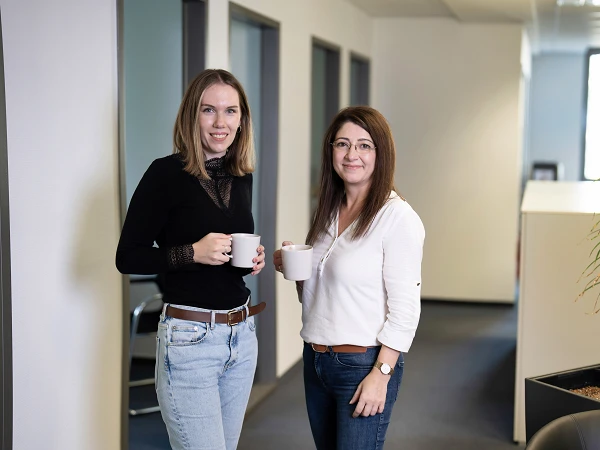 Zwei Frauen stehen in einem modernen Büroumfeld auf einem Flur und halten jeweils eine Tasse in der Hand. Die Frau links trägt ein schwarzes Oberteil und Jeans, während die Frau rechts ein weißes Oberteil und eine braune Hose trägt. Im Hintergrund sind Bürotüren und ein Pflanzenkübel sichtbar.